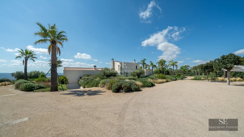 View of infinity pool surrounded by a modern villa with large windows and minimalist design. Well-manicured garden and blue sky.