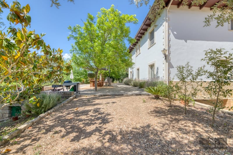 Sunny gravel garden with mature shade trees, outdoor dining set, and white Mediterranean house facade.