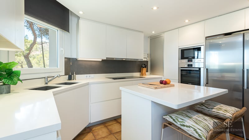 Bright modern kitchen featuring white cabinets, stainless steel appliances, a center island, and a window with a forest view.