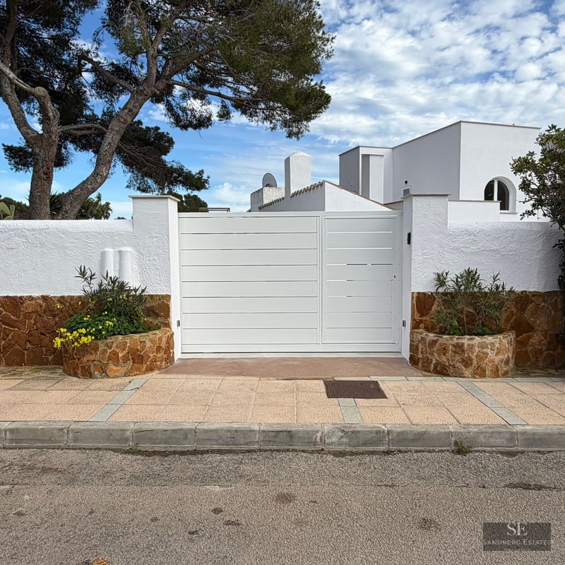 Large white metal gate flanked by white stucco and brown stone walls under a blue sky with a pine tree.