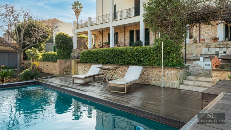 Turquoise swimming pool with dark wood deck, sun loungers, and a rustic stone villa in the background.
