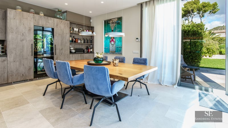Salle à manger moderne avec table en bois, chaises bleues, cave à vin et grande baie vitrée ouvrant sur le jardin.