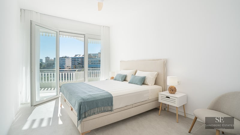 Bright bedroom with a double bed, bedside tables, and large glass doors opening to a balcony with a city view.