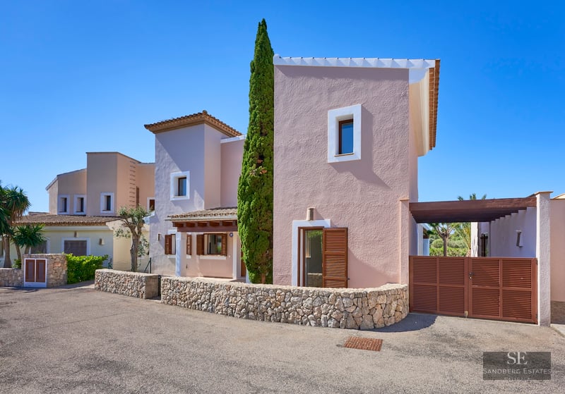 A salmon-colored Mediterranean villa with a tall cypress tree, a low dry stone wall, and a bright blue sky.