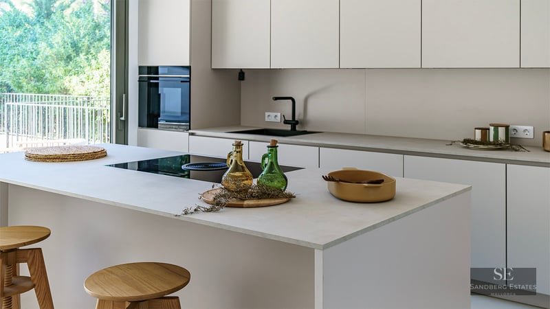 Clean minimalist white kitchen featuring a large island, wooden stools, and views to a lush garden through glass doors.