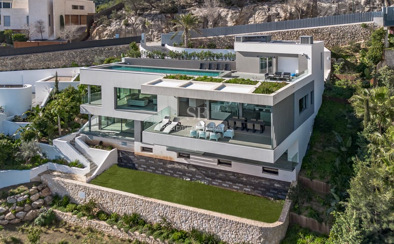Vue aérienne d'une villa moderne blanche à plusieurs niveaux avec une piscine à débordement sur le toit.