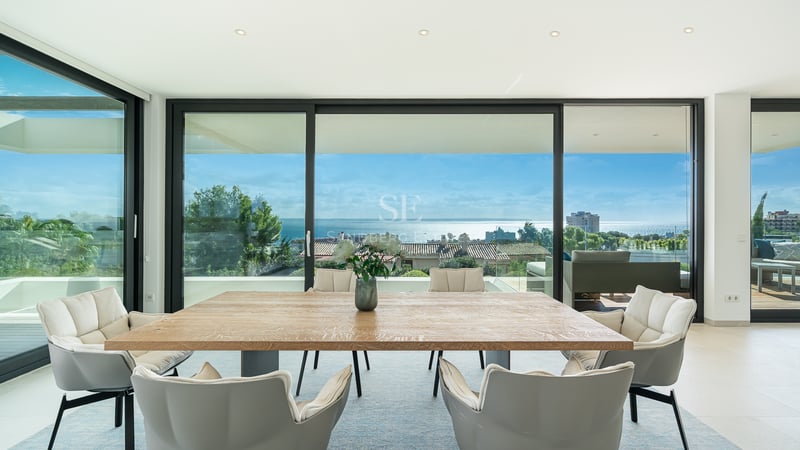Salle à manger lumineuse avec table en bois et chaises grises face à des baies vitrées avec vue sur l'océan.