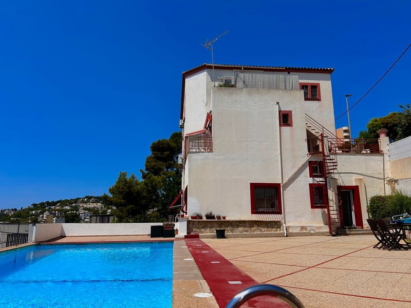 Large outdoor swimming pool in front of a white villa with red trim under a bright blue sky.