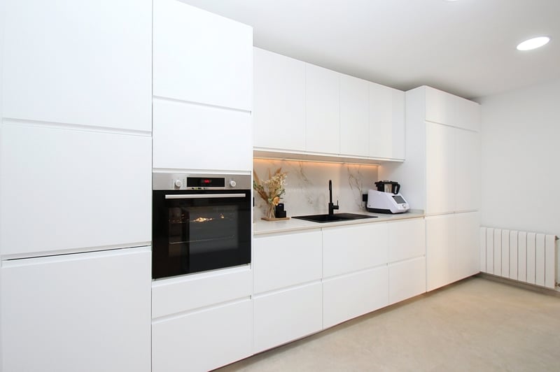 Modernt vitt kök med handtagsfria skåp, svart kran och integrerad ugn i rostfritt stål.