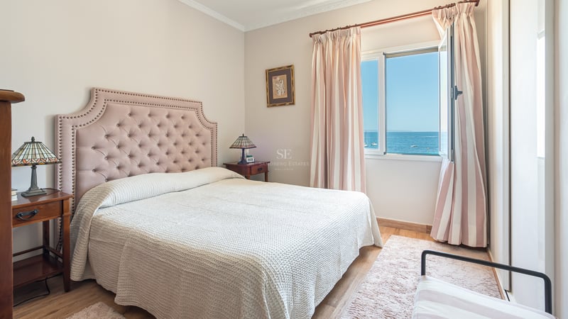 Ljust sovrum med rosa stoppad huvudgavel, trämöbler och fönster som öppnar upp mot det blå Medelhavet.