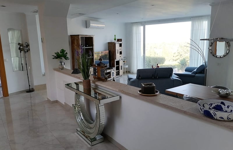 Salon moderne avec sol en marbre, canapés bleus et grande baie vitrée donnant sur un jardin verdoyant.