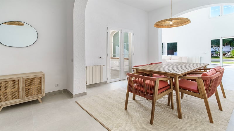 Ljus matsal med träbord, stolar med terrakottafärgade dynor, flätad taklampa och vita valv.