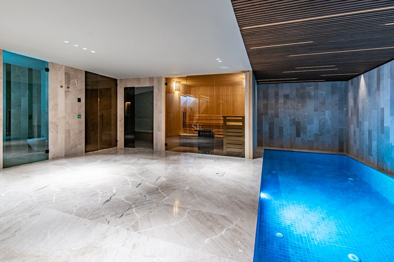 Spa intérieur avec petite piscine bleue, sauna vitré et sol en marbre sous un plafond à lattes.