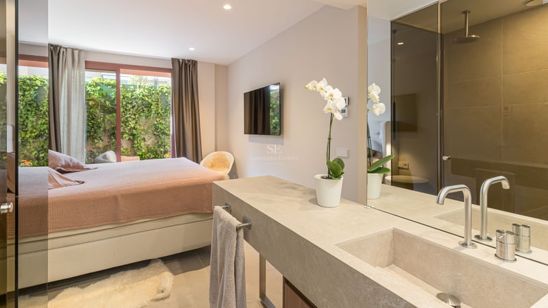 A modern master bedroom featuring a natural stone vanity and sink, a large mirror, and access to a green patio.