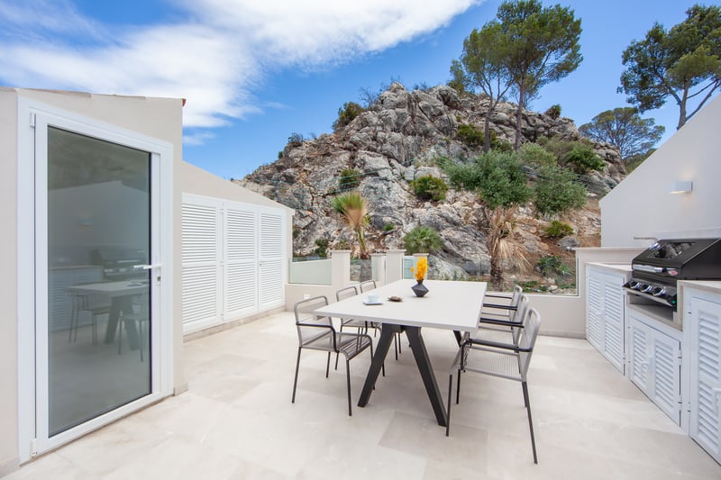 Terrasse blanche moderne avec table à manger, chaises et barbecue, avec une montagne rocheuse en arrière-plan.
