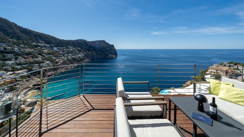 Elevated wooden terrace with lounge seating overlooking a Mediterranean bay and rugged coastline under a blue sky.