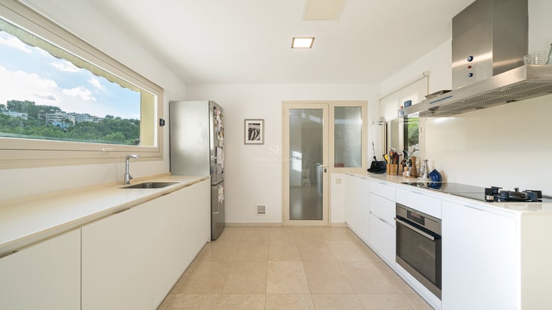 Ljust kök med vita skåp, rostfri kyl och ugn, samt ett stort fönster med utsikt över grönska.