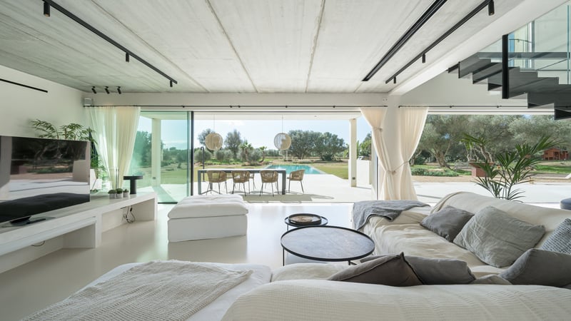 Ljust vardagsrum med minimalistisk inredning och stora glasdörrar mot en utomhuspool och matplats.