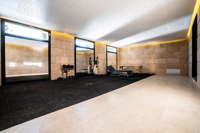 Fitnessraum mit Natursteinwänden, schwarzem Gummiboden, Massageliege und indirekter Deckenbeleuchtung.