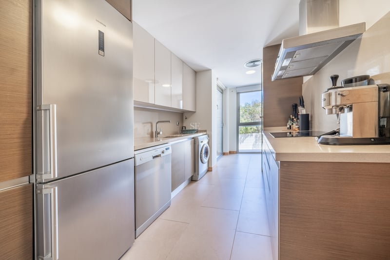 Cuisine en couloir avec placards en bois clair, appareils en inox et grande fenêtre au bout.