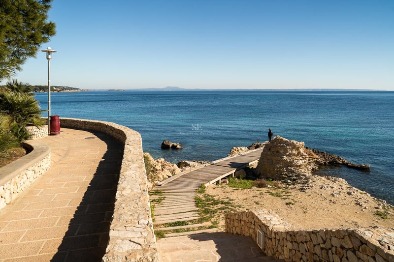 Küstenpromenade aus Naturstein und Holzsteg zum türkisfarbenen Mittelmeer unter blauem Himmel.