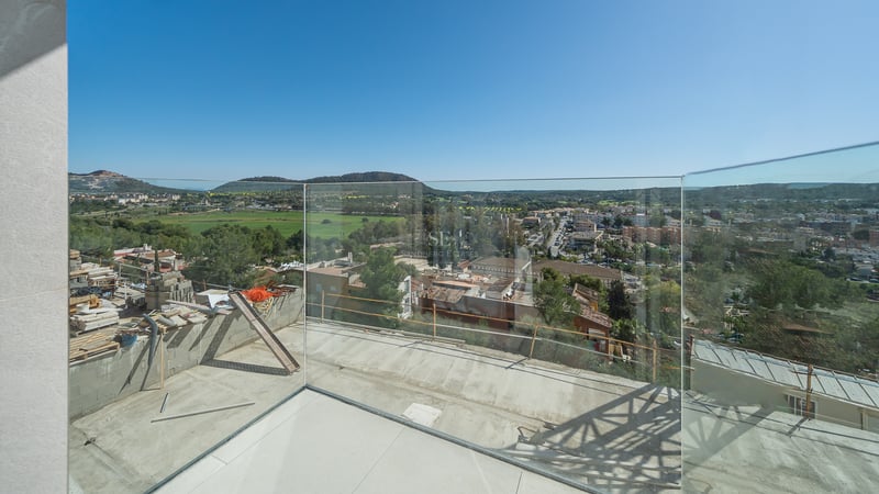 Terrasse moderne en construction avec garde-corps en verre surplombant un village méditerranéen et des collines.