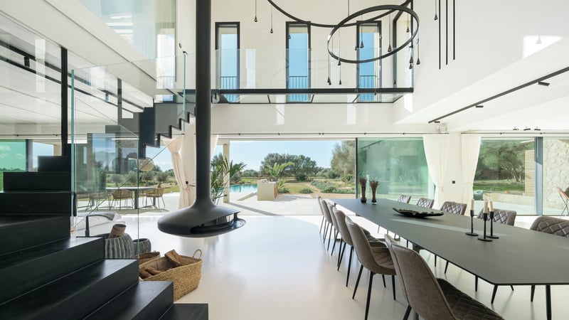 Ljus matsal med dubbel takhöjd, svart hängande öppen spis, glasräcken och utsikt över trädgård och pool.
