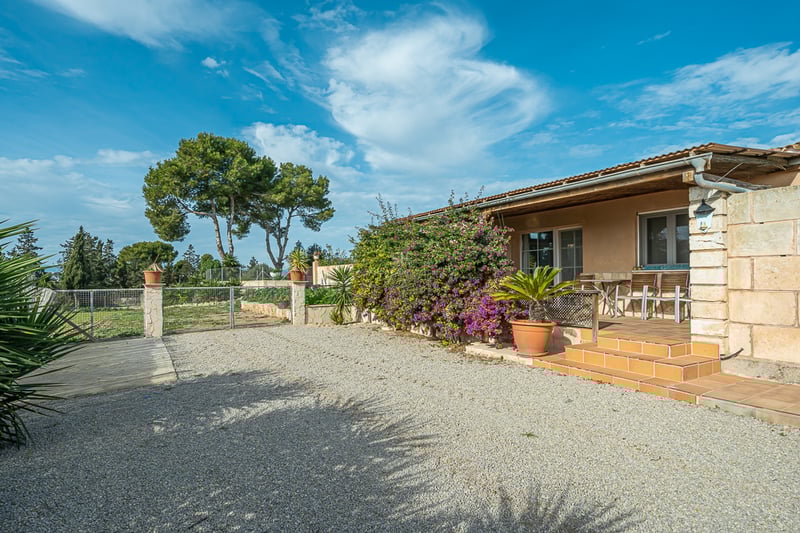 Façade en pierre d'une villa avec porche en terre cuite, cour en gravier et bougainvilliers sous un ciel bleu.