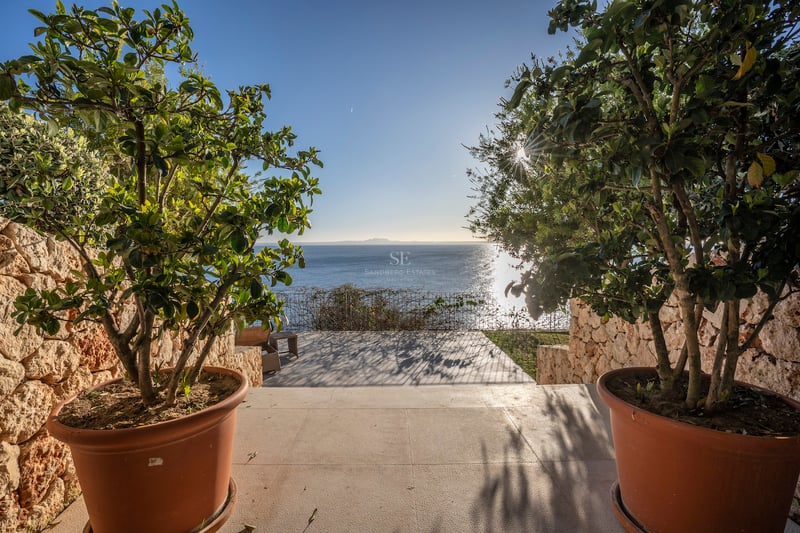 Blick auf das blaue Meer von einer Steinterrasse mit Topfpflanzen und Natursteinmauern unter blauem Himmel.