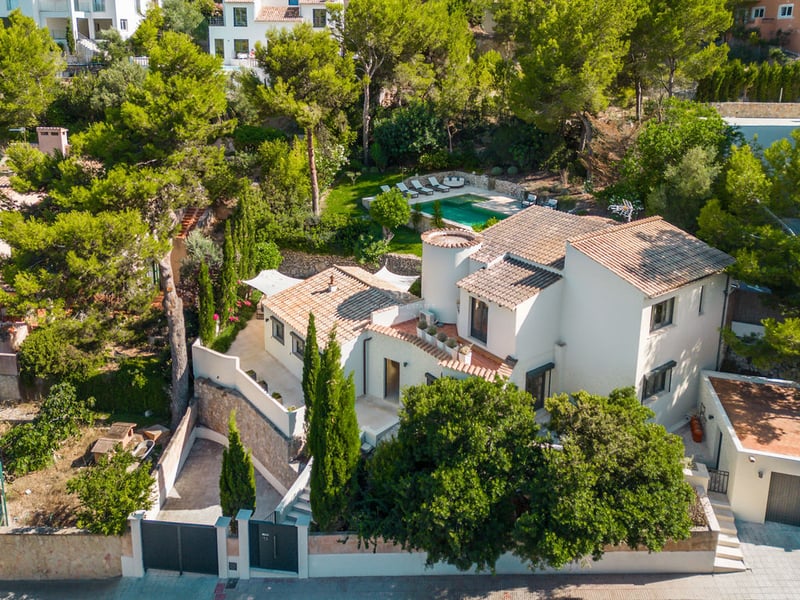 Aerial view of a white multi-level villa with terracotta roof and pool surrounded by lush pine trees.