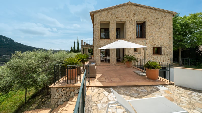 Villa en pierre naturelle avec grande terrasse en terre cuite, parasol blanc et salon de jardin sous un ciel bleu.