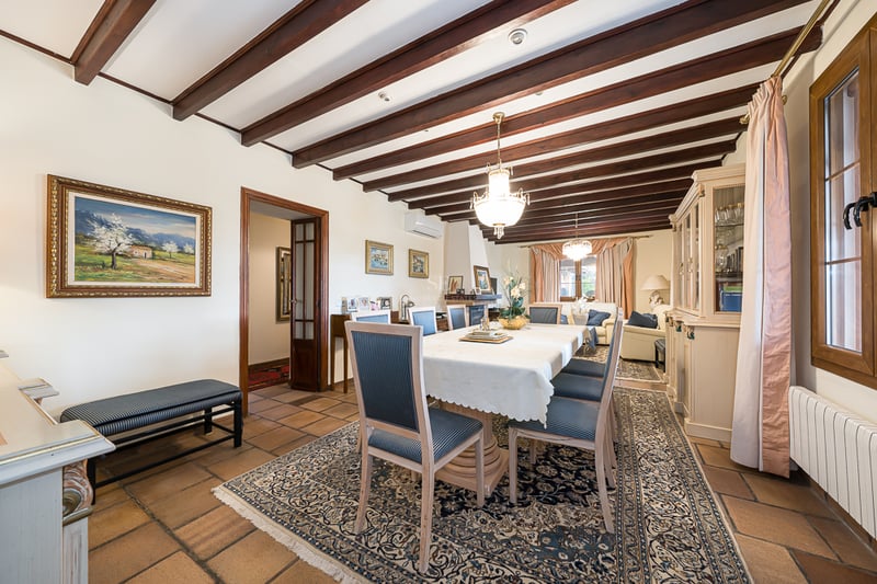 Salle à manger formelle avec poutres en bois sombre, sol en terre cuite et grande table sous un lustre classique.