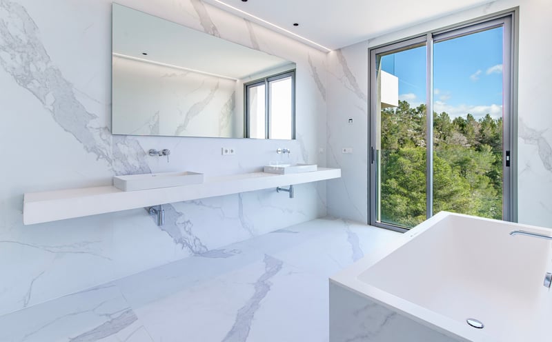 Salle de bain principale lumineuse avec murs et sols en marbre blanc, double vasque, baignoire et vue sur la forêt.