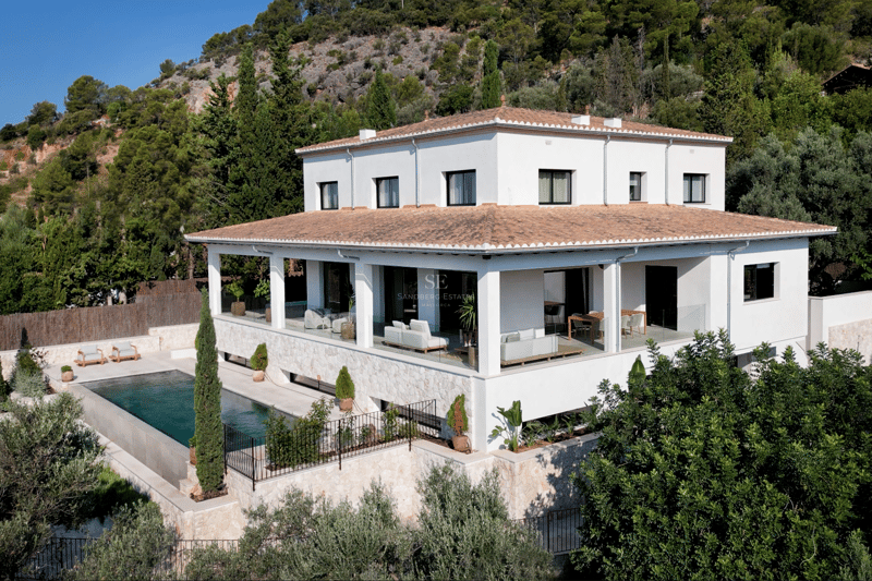 Elevated view of a white multi-level villa with a terracotta roof, infinity pool, and stone-clad terrace in a lush setting.