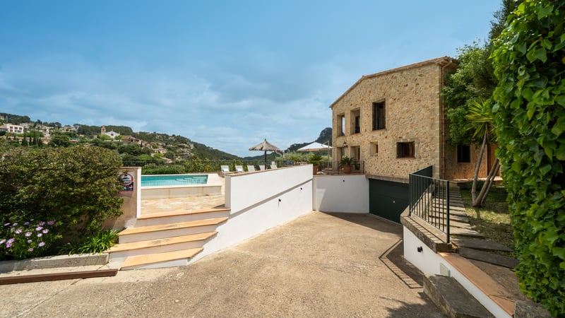Vue extérieure d'une villa en pierre avec piscine, terrasse ensoleillée et vue sur la montagne par temps clair.
