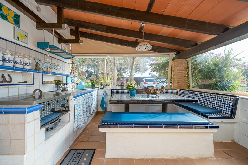Terrasse couverte avec cuisine d'été en carreaux bleus et blancs, bancs intégrés et plafond à poutres en bois.