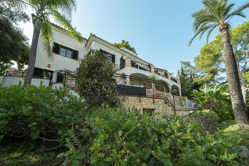 Vit trevåningsvilla med valvterrasser, stenmurar och palmer under en klarblå himmel.