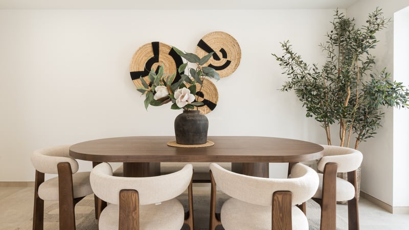 Dining room featuring an oval wooden table, cream chairs, woven wall art, and a large indoor olive tree.