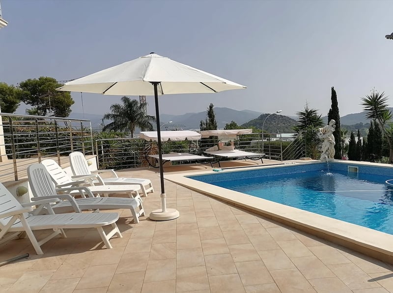 Espace piscine ensoleillé avec chaises longues blanches, parasol et statue décorative surplombant les montagnes.