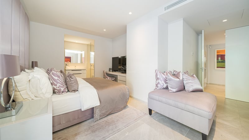 Modernes Schlafzimmer mit Doppelbett, mauvefarbener Recamiere, Schreibtisch und Blick ins Badezimmer.