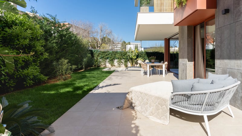 Modern terrace with stone tiling, outdoor dining set, designer lounge chair, and a manicured green lawn under a blue sky.