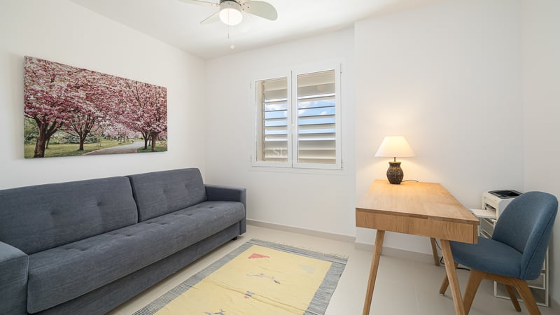 A minimalist room with a grey sofa, wooden desk with a lamp, a blue chair, and a window with white shutters.
