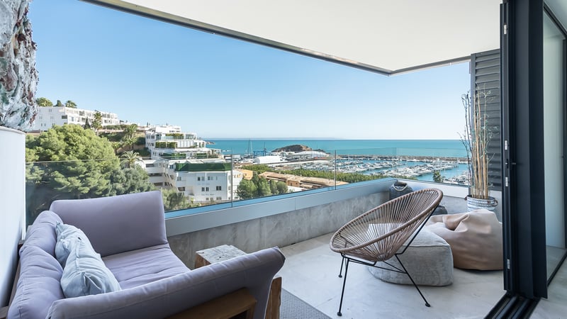 Überdachte Terrasse mit lavendelfarbenem Sofa, Korbstuhl und Glasgeländer mit Blick auf den Yachthafen und das Meer.