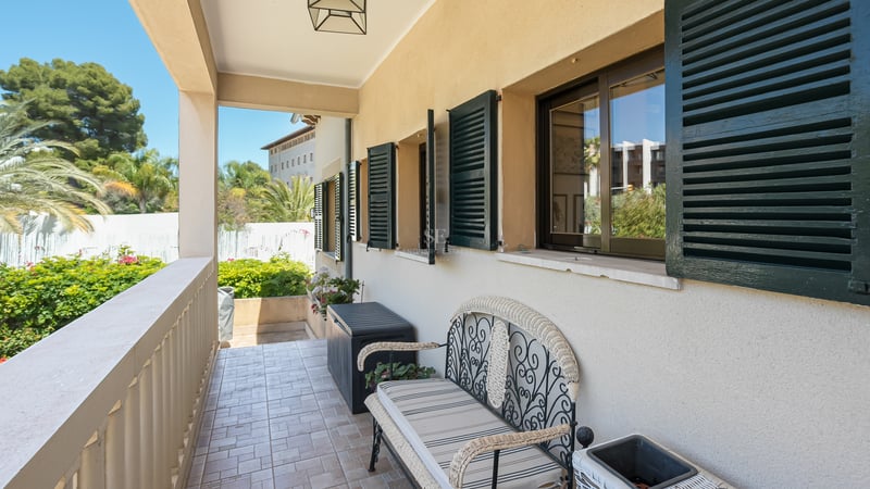 Täckt balkong med smidesbänk, gröna fönsterluckor och utsikt över en grönskande trädgård.