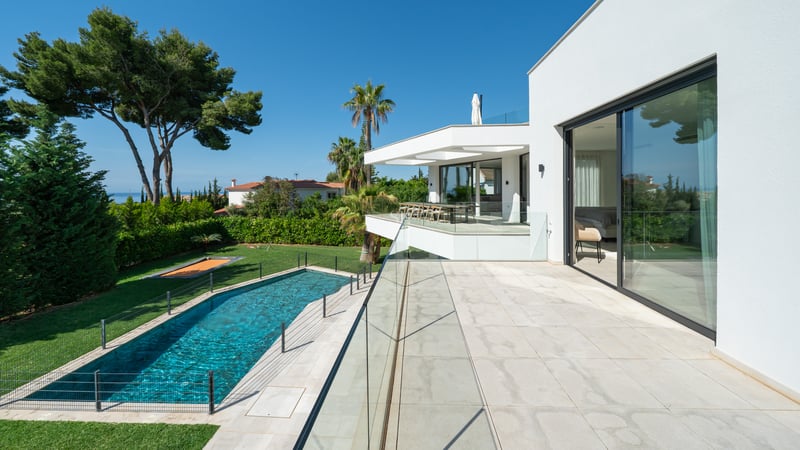 Vue surélevée depuis une terrasse moderne blanche sur une piscine rectangulaire, une pelouse et des pins.