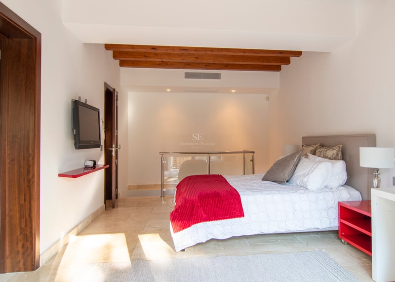 Modernt sovrum med dubbelsäng, röda detaljer, synliga träbjälkar och ljust stengolv.