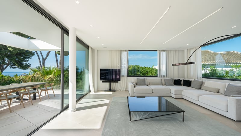 Salon moderne et lumineux avec de grandes baies vitrées ouvrant sur une terrasse avec vue mer et un canapé d'angle blanc.