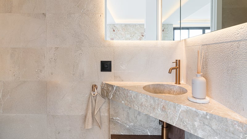 Nahaufnahme eines integrierten Natursteinwaschbeckens mit gebürsteter Messingarmatur und hinterleuchtetem Spiegel.