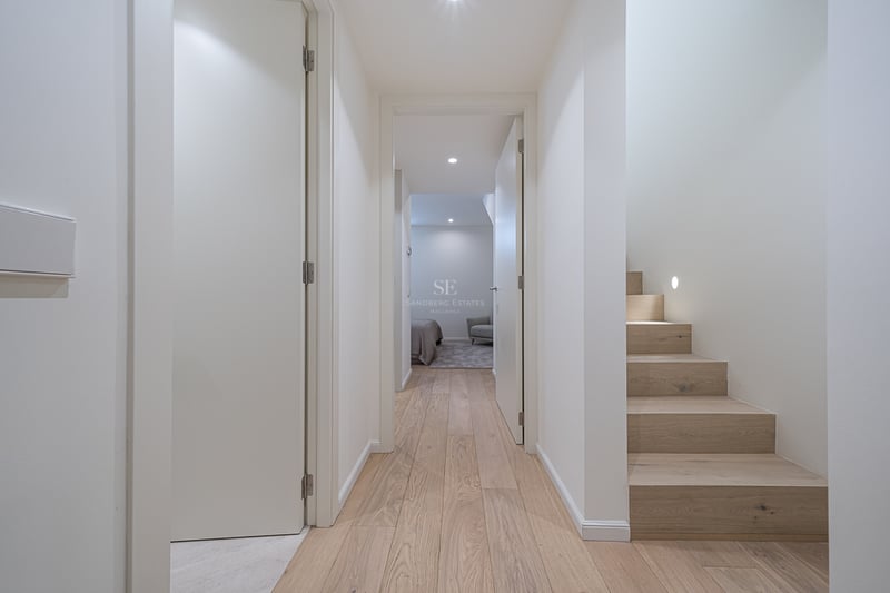 Bright modern hallway with light wood floors, white walls, and a wooden staircase with integrated wall lighting.
