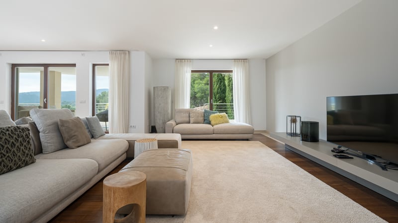 Salon spacieux avec canapés beiges, pouf en cuir, parquet sombre et grandes fenêtres donnant sur des collines verdoyantes.